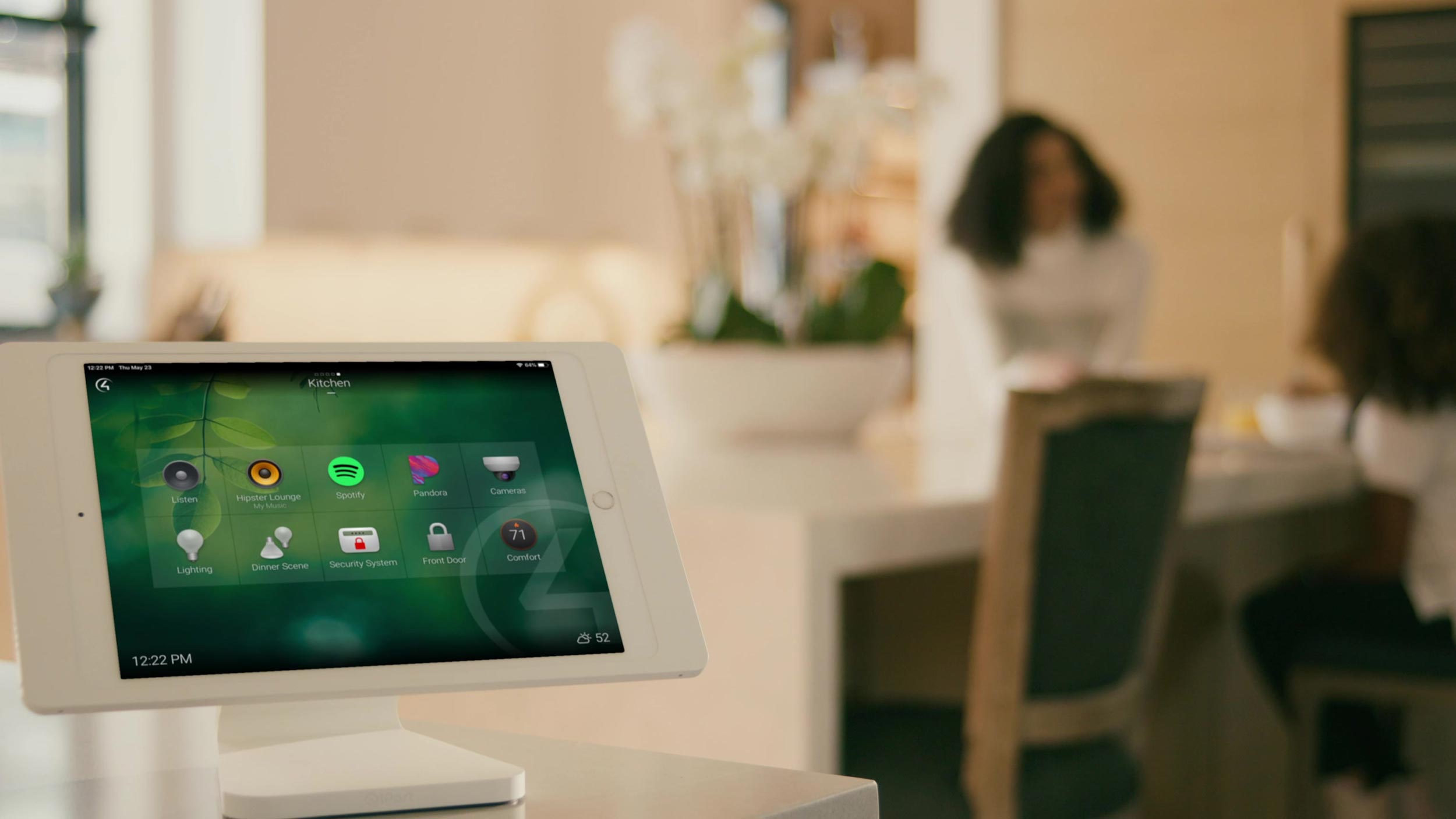 Smart home tablet on a countertop displaying a control dashboard—icons for music (Spotify, Pandora), lighting, dinner scene, security system, front door, cameras, and thermostat—set in a bright kitchen with two people softly blurred in the background.